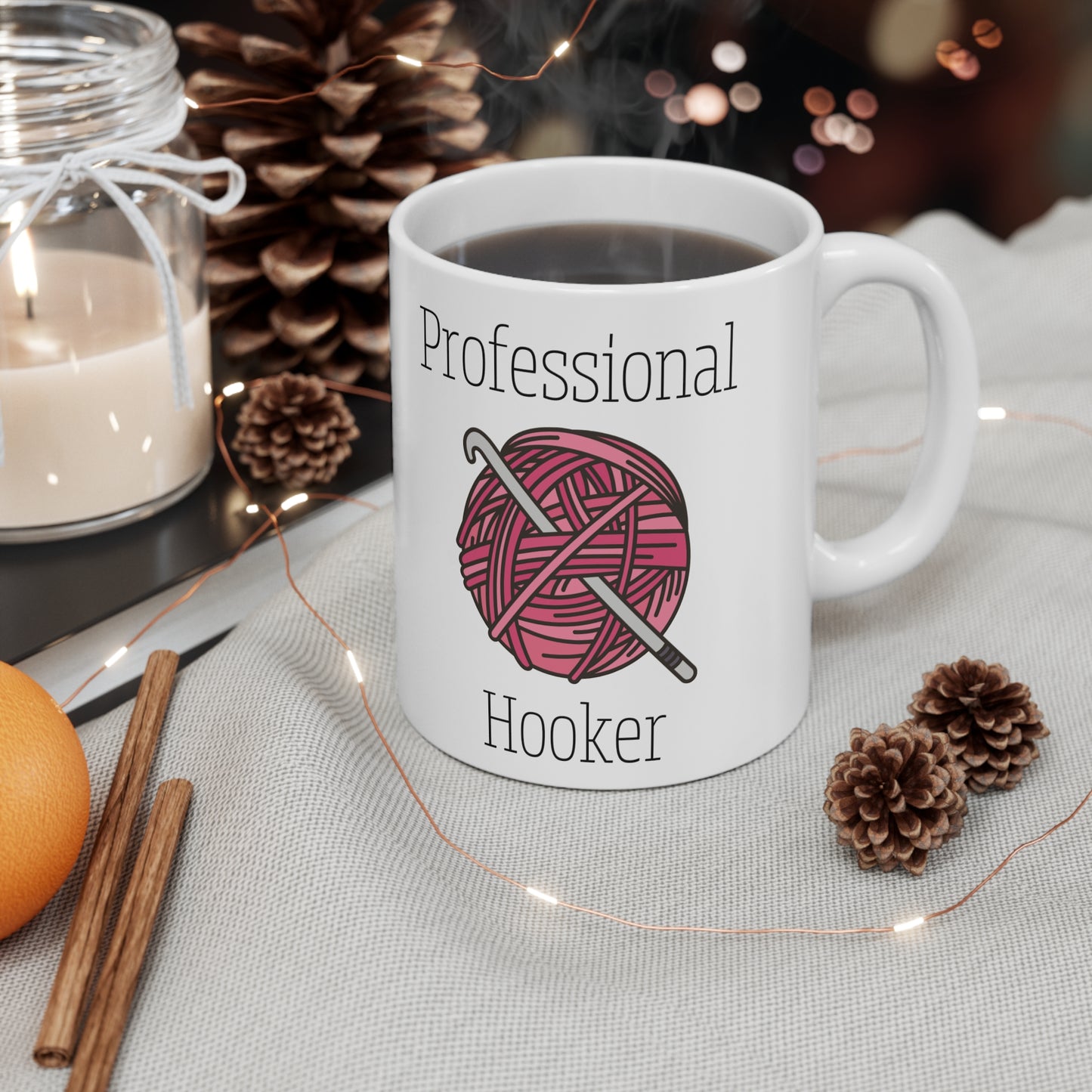 White mug with 'Professional Hooker' design featuring yarn and a hook, placed on a table with decorative items.